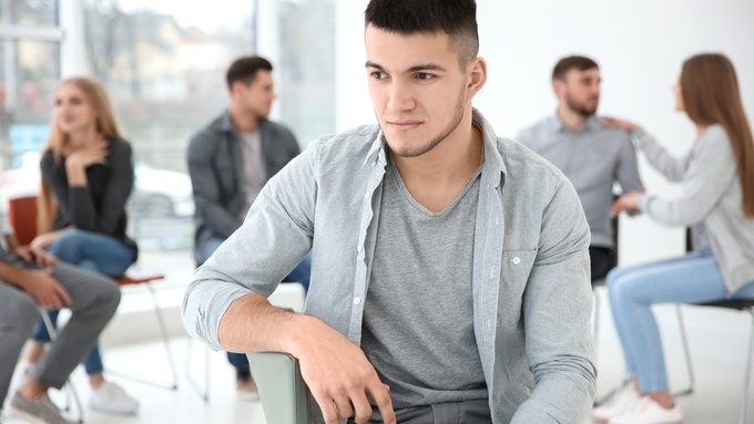 A man sits sideways in a chair facing away from a group of people sitting in a circle talking to each other.