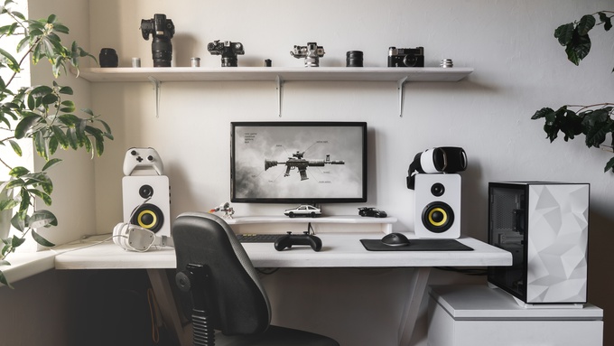 A gaming desk set up with speakers, a VR headset, a monitor, and controllers. A shelf of cameras is above the desk.