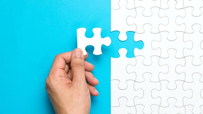 A close-up view shows a hand holding a plain white puzzle piece next to a puzzle. The background is blue.
