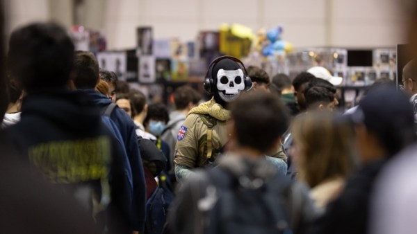 A man wearing a skull mask, headphones, and a green military costume stands in the middle of a blurred crowd at a convention.
