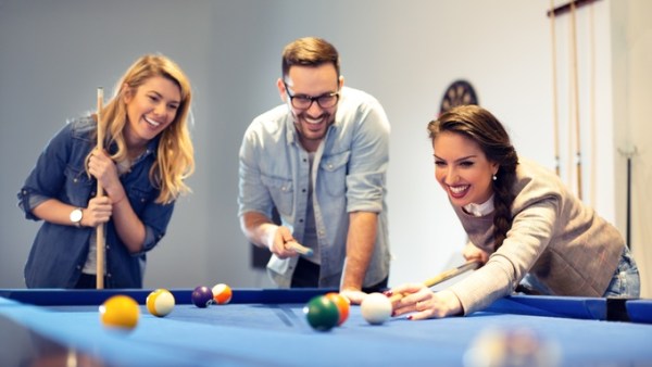 Two women and one man holding pool cues while playing pool on a blue table. One woman is bending over to hit a ball.