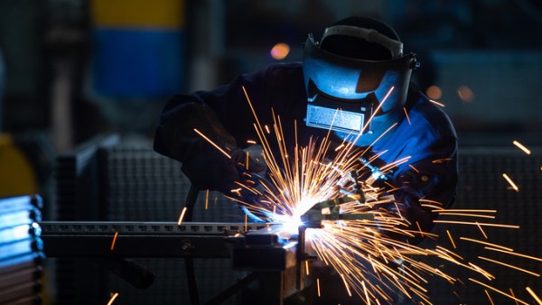 A welder wearing a welding helmet leans forward as they work on a piece of steel. Orange sparks fly in all directions.