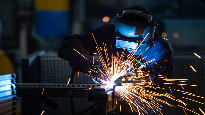 A welder wearing a welding helmet leans forward as they work on a piece of steel. Orange sparks fly in all directions.