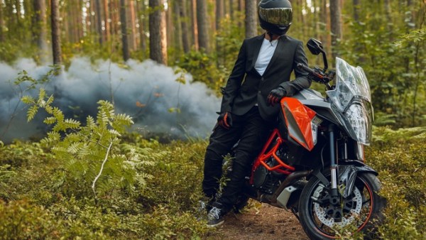 A person wearing a black suit, helmet, and sneakers leaning against a black-and-orange motorbike in the woods.