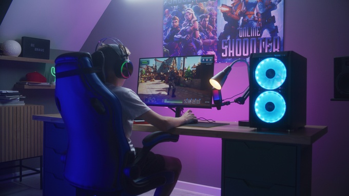 A gamer sitting at their PC gaming desk in a dimly lit room. Two video game posters hang on the wall in front of the desk.