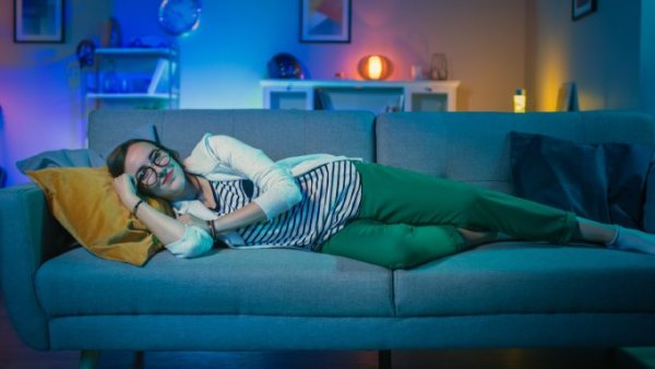 A woman is lying on a sofa in a living room with multicolored lighting and framed artwork on the walls.