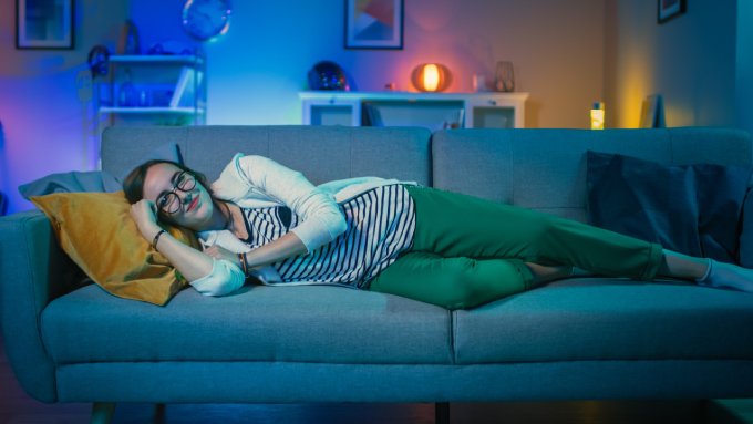 A woman is lying on a sofa in a living room with multicolored lighting and framed artwork on the walls.