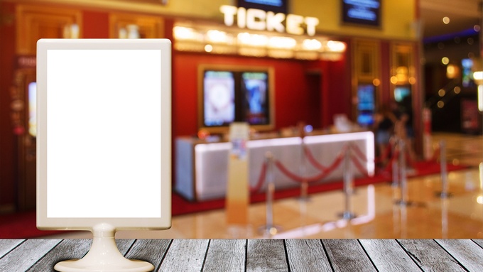 A blank white sign sits on a wooden table. In the background is a view of the ticket sales counter at a movie theater.