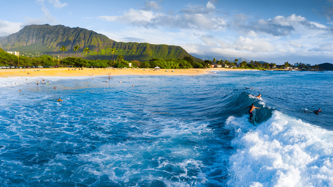 Surfers ride an ocean wave at a popular surf spot in Oahu, Hawaii. Other people swim in the shallow water along the coast.