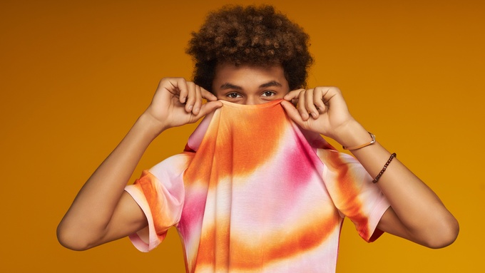 A person lifting the collar of their tie-dye T-shirt to shyly hide the lower half of their face from the viewer.