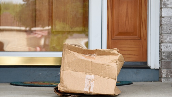 A damaged brown box placed in front of a brown door, and a welcome mat on the floor outside during the day.