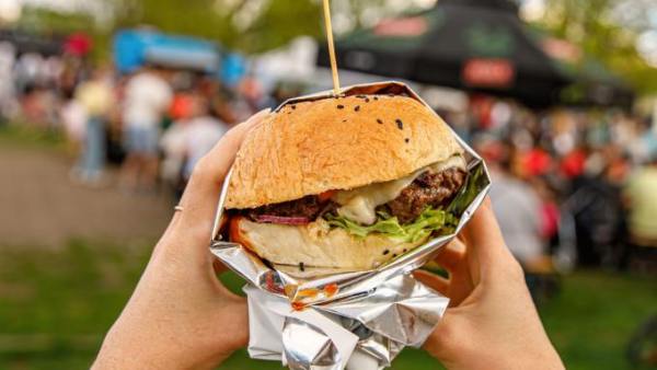 A close-up view shows a person wearing a black watch holding a burger wrapped in aluminum foil. The background is blurry.