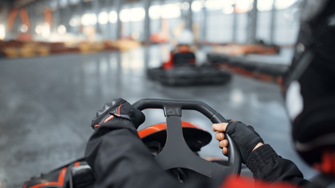 A point-of-view shot of a driver with their hands on the steering wheel, maneuvering a go-kart through an indoor area.
