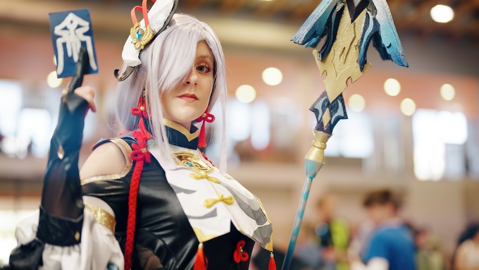 A female wearing a cosplay outfit holds a card in one hand and a multi-colored metal weapon in her other hand.