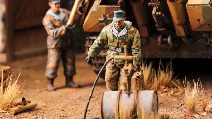 A close-up of a historical diorama. Two male soldiers are carrying a toy gas can next to large tanks.