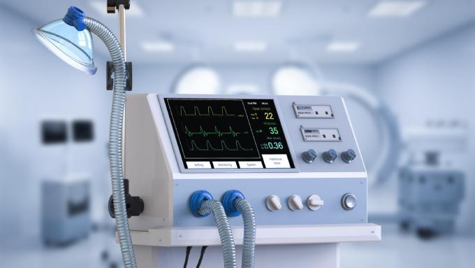 A close-up of a medical ventilator with a digital screen showing data, attached hoses, and a breathing mask.