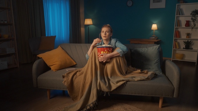 A woman sits on a couch, eating popcorn while watching something. She has a shocked expression on her face.