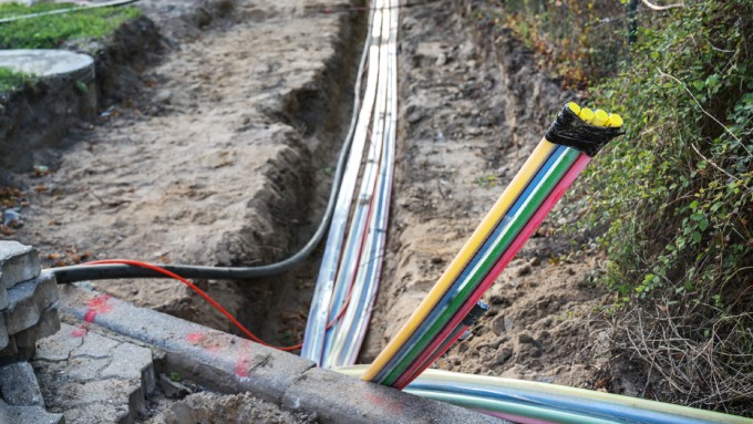 A muddy trench aligns with an urban street and houses a collection of fiber-optic cables. A fence lines the trench.