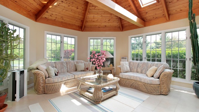 An open sunroom features two beige sofas, large windows, a vaulted ceiling, a striped rug, and a beige tiled floor.