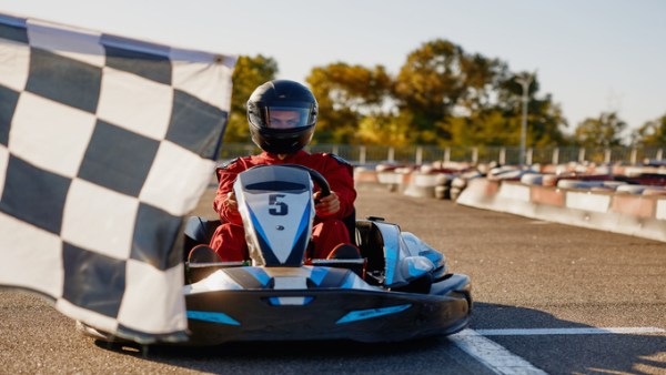 A person wearing a red jumpsuit and a black helmet sits in a black go-kart on an outdoor track. A checkered flag is in front.