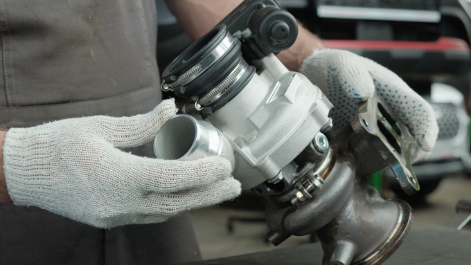 Gloved hands hold a turbocharger with bolts, clamps, and a black coupling, set in a blurred workshop background.