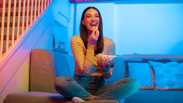 A woman relaxes on a couch with popcorn during movie night, the room lit by vibrant blue and pink smart home lighting.