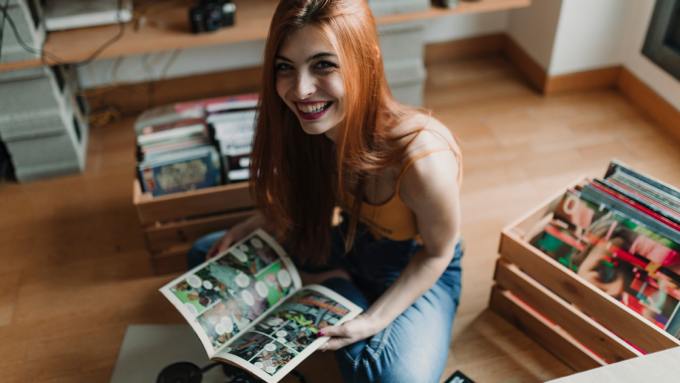 A young woman sits on the floor happily with her collection of comic books in a wooden crate next to her.