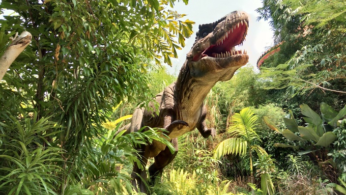 A massive roaring Tyrannosaurus Rex stands in a lush green jungle surrounded by tropical plants and trees.