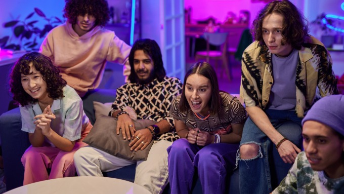 Group of friends sit on couch playing video game together, reacting excitedly in neon-lit living room.