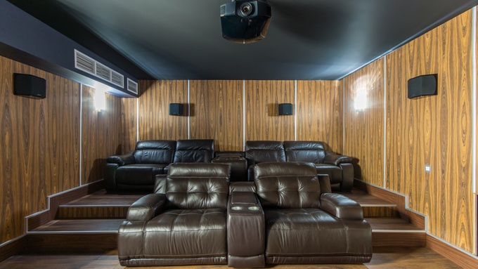 A well-lit room with wooden walls features large brown leather recliners, speakers, and a black projector.