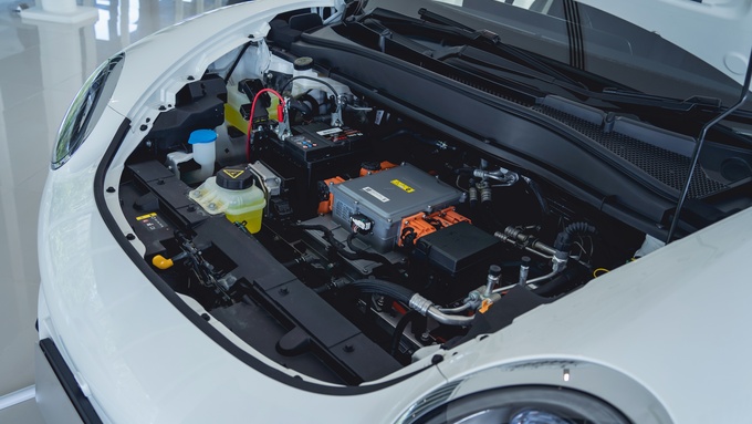 A white electric vehicle parked on a white floor. The EV hood is open to reveal the battery and components inside.