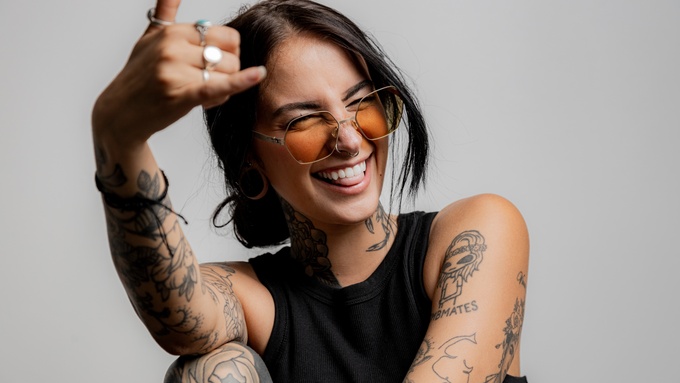 A young person with tattoos on both arms sticking their tongue out and making a "love" sign with their hand.