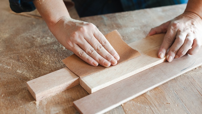 A pair of hands using a small bit of sandpaper to sand away at a thin plank of wood, sending sawdust into the air.
