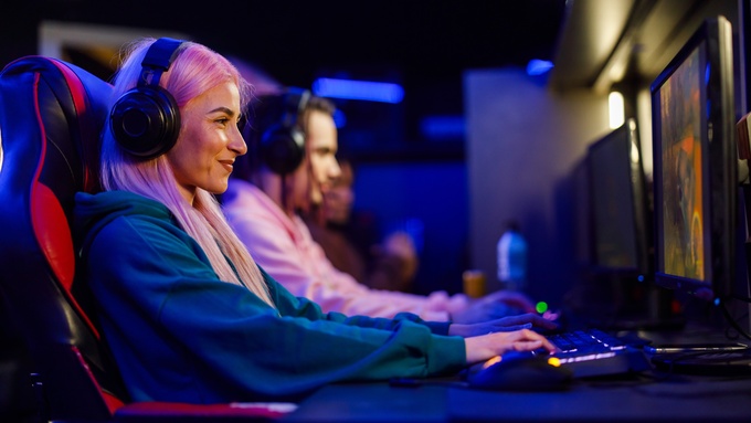 A woman with pink hair wearing a headset plays a video game at a neon-lit gaming station beside other players.