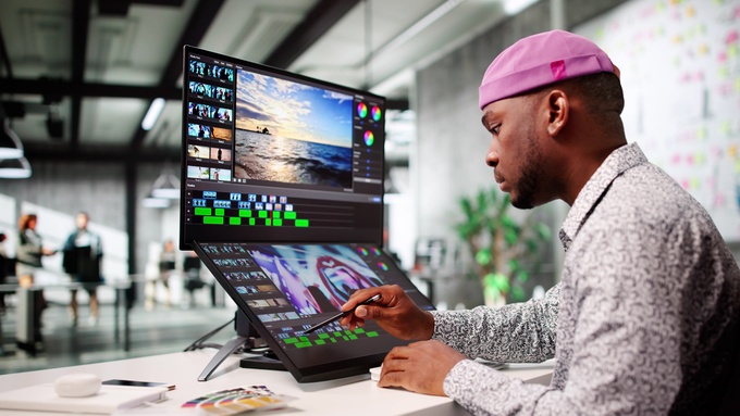 Video editor wearing pink cap uses stylus on touchscreen monitor with timeline and color tools in studio.