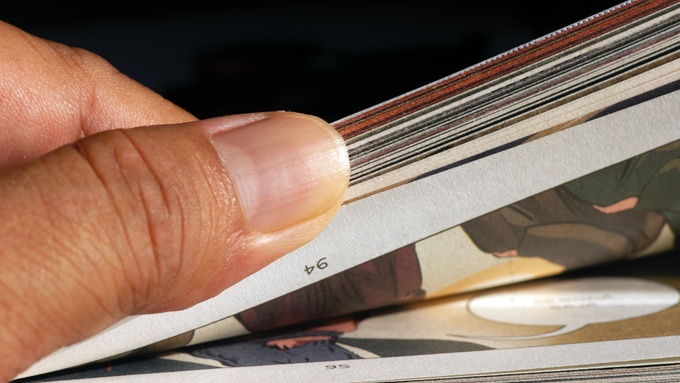 A hand flips through a stack of comic book pages, showing the layered edges and printed artwork inside.
