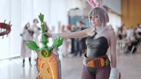 A cosplayer wearing a black outfit stands in a busy convention hall and holds a large orange and green sword prop.