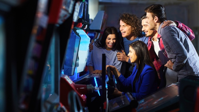 A group of young adults around an arcade racing game, smiling and laughing as one drives while the others cheer.