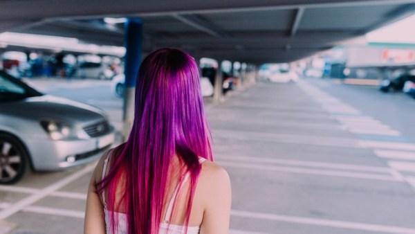 A person walks through a parking garage full of cars. They have long, purple and magenta straight hair.
