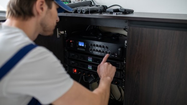 A technician wearing a blue hat and crouching over to adjust an audio system inside a wooden cabinet.