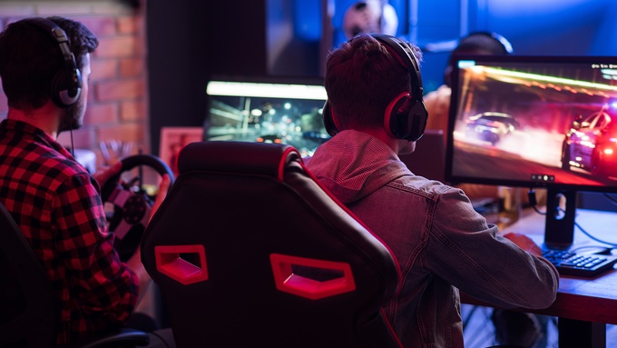 Two gamers wearing headsets play a racing video game on their own monitors at a desk in a dimly lit room.