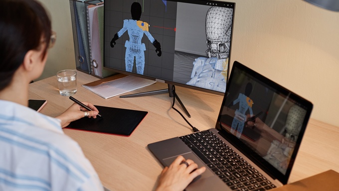 A person sits at a desk with a laptop, a monitor, and a digital drawing pad. The outline of a person is on the screens.