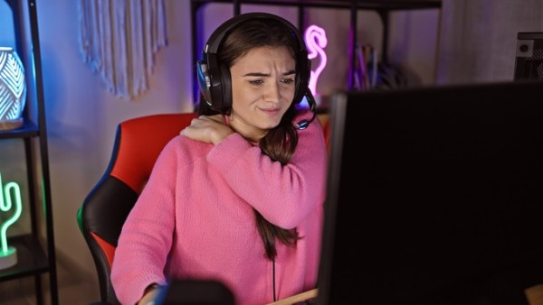 A gamer rubs her shoulder while sitting in her gaming room at night. She is wearing a pink sweatshirt.