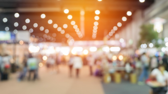 A blurry view of a convention hall with people walking around to visit booths and interact with one another.