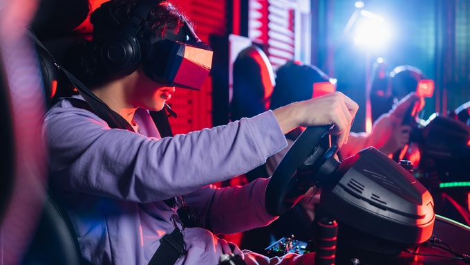 A teenager wearing a VR headset and holding a racing simulator steering wheel in a neon-lit indoor game area.