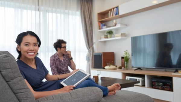 Two adults sitting on the sofa while one holds a tablet that has smart home controls on it. The TV in the room is off.