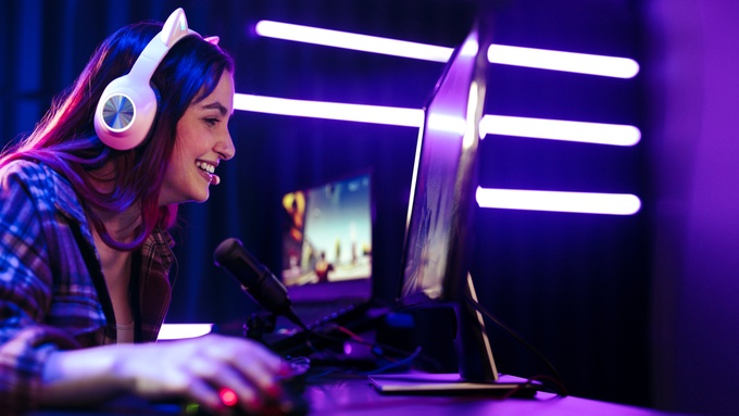 A young female streamer at her computer desk wears a headset and smiles at the monitor. The lights are purple.