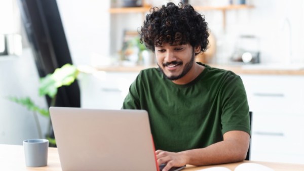 Man with curly hair works on a laptop at a wooden table beside a gray mug and open book in a sunlit room.