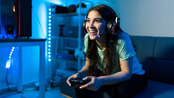 In a blue-lit room, a woman on a couch wears a headset and holds a gaming controller. A computer tower is beside her.