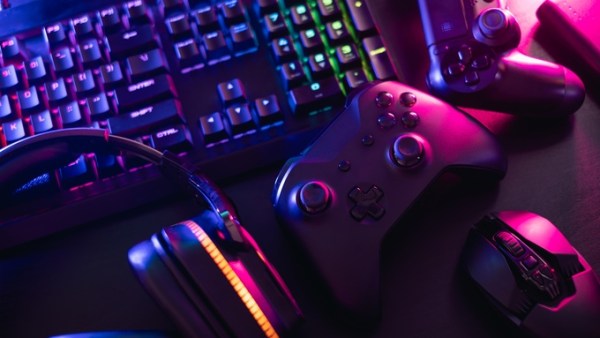 A close-up view shows two gaming controllers, a computer mouse, a gaming headset, and a desktop keyboard on a flat surface.